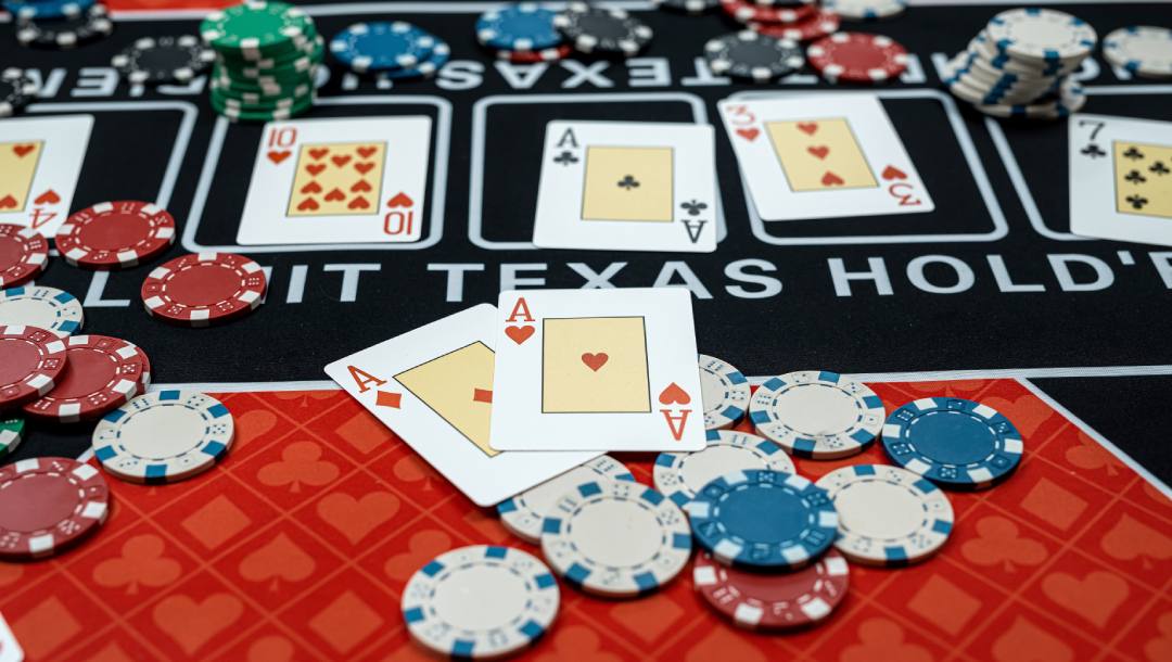 A photograph of a Texas Hold’em table, showing two Ace playing cards on top of scattered poker chips at one of the table positions, with five face-up cards in the center of the table.