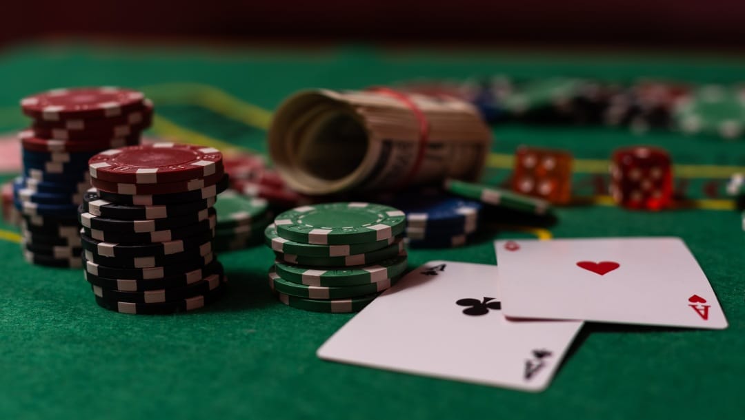 A close-up photograph of two ace playing cards, face-up on a poker table, surrounded by chips, dice, and rolled up cash.