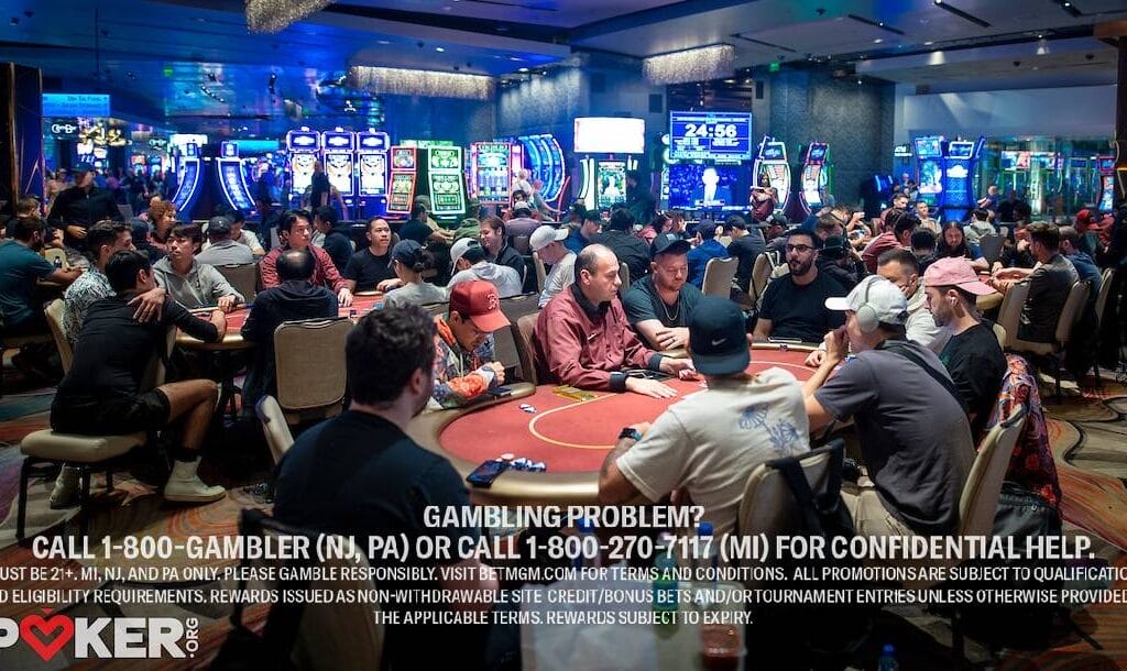 People are sitting around tables playing poker in a casino with bright slot machines in the background.