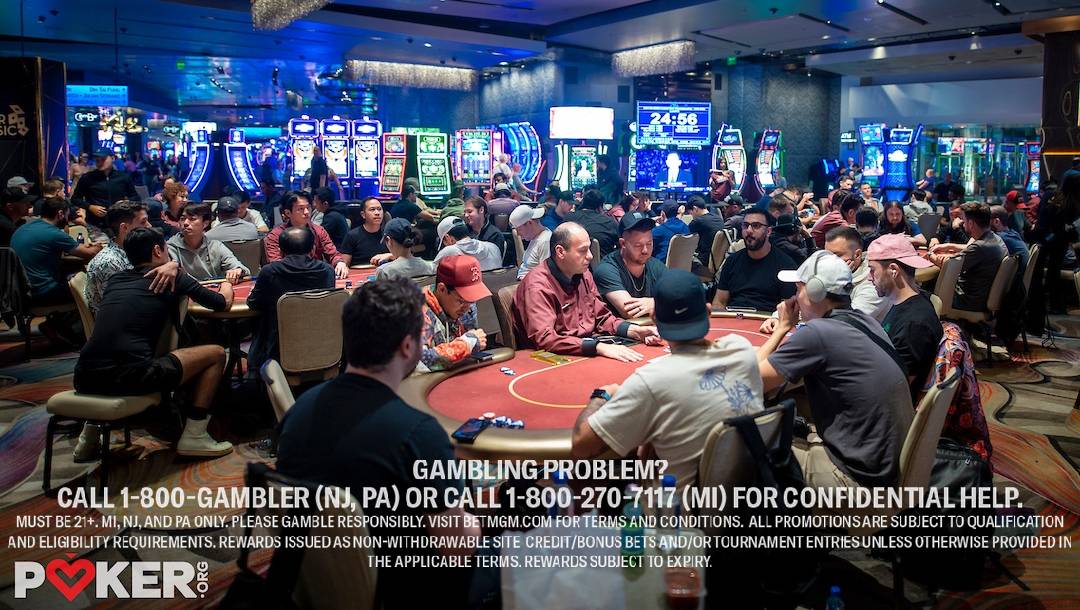 People are sitting around tables playing poker in a casino with bright slot machines in the background.
