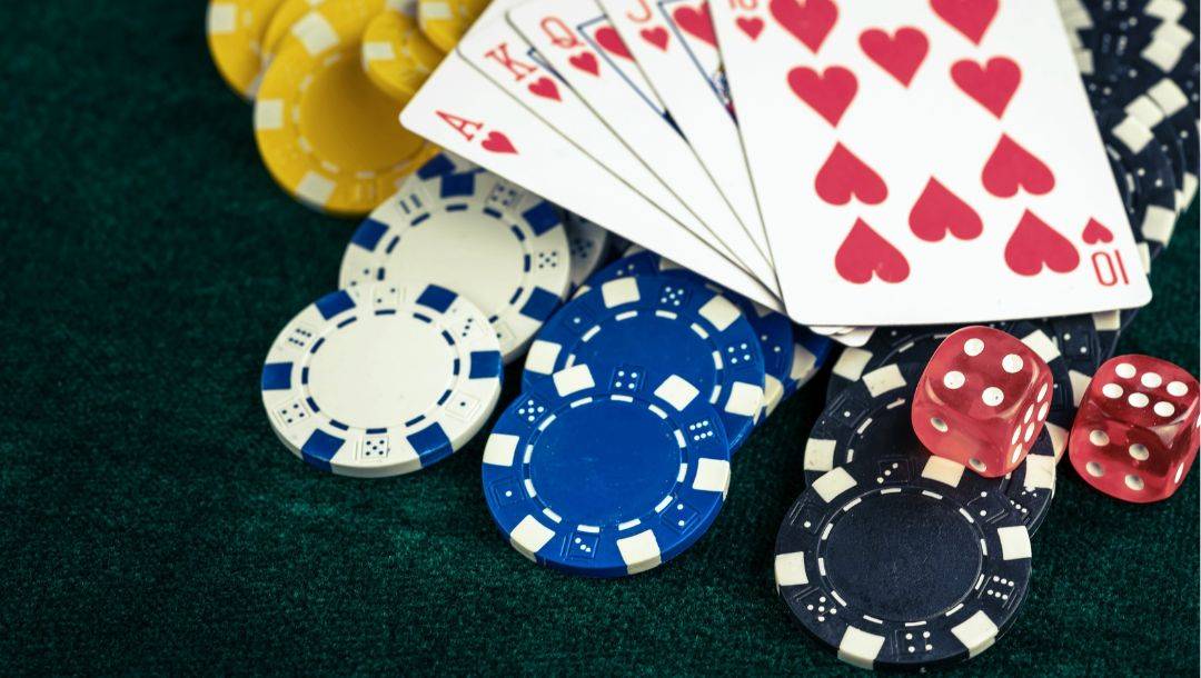 An Ace of Hearts, King of Hearts, Queen of Hearts, Jack of Hearts, and 10 of Hearts arranged in a royal flush on a poker table, atop stacks of poker chips, with two red dice