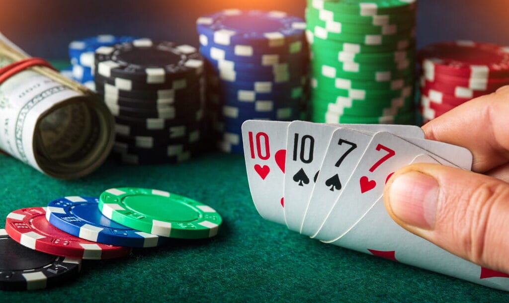 A person seated at a poker table, revealing a hand of cards: a 10 of Hearts, a 10 of Spades, a 7 of Spades, and a 7 of Hearts. The table is adorned with a roll of cash and stacks of poker chips.
