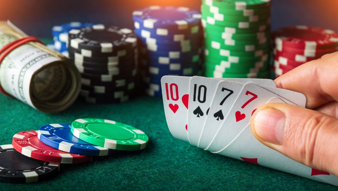 A person seated at a poker table, revealing a hand of cards: a 10 of Hearts, a 10 of Spades, a 7 of Spades, and a 7 of Hearts. The table is adorned with a roll of cash and stacks of poker chips.