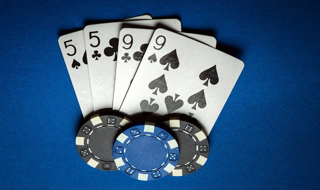 A five of spades, a five of clubs, a nine of spades, and a nice of clubs, arranged on a blue table, featuring two black casino chips, and one blue casino chip.