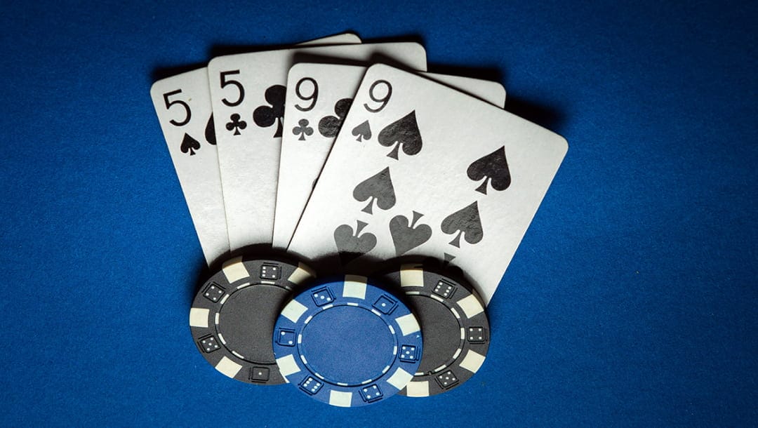 A five of spades, a five of clubs, a nine of spades, and a nice of clubs, arranged on a blue table, featuring two black casino chips, and one blue casino chip.