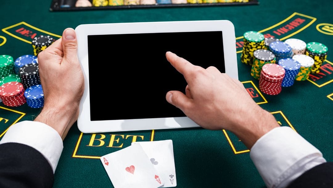 A first-person view of a person holding a tablet while resting their arms on a casino poker table with several stacks of poker chips next to him.