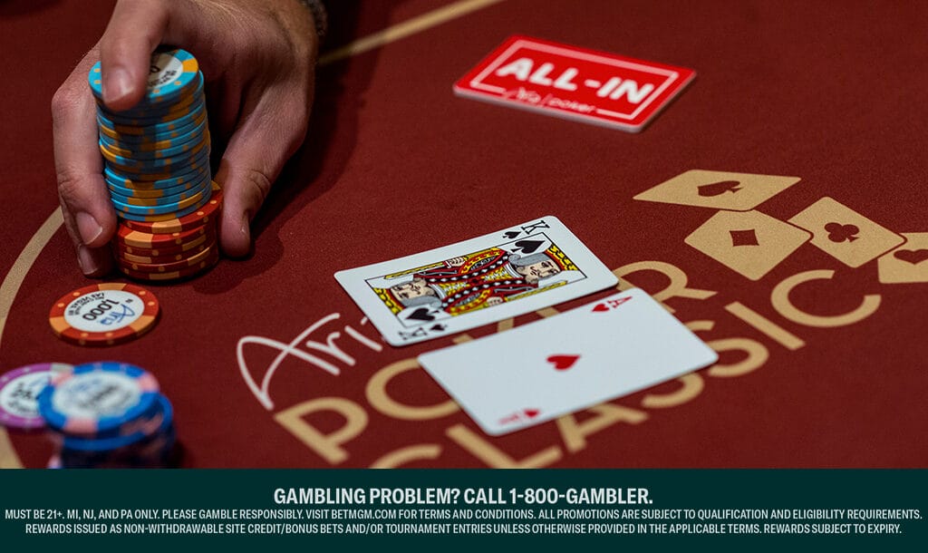 A poker player holds a large stack of different colored poker chips between their fingers. There are smaller stacks of poker chips next to their hand. A king and an ace sit in the center of a dark red poker table.