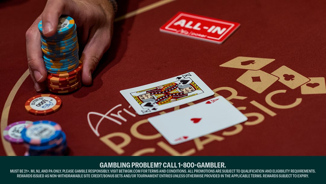 A poker player holds a large stack of different colored poker chips between their fingers. There are smaller stacks of poker chips next to their hand. A king and an ace sit in the center of a dark red poker table.