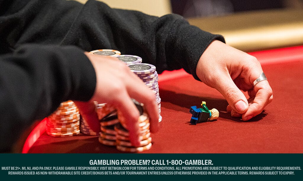 A poker player shuffles two stacks of poker chips with their one hand while their other hand rests on a red poker table next to a Lego figure.