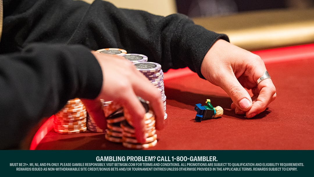 A poker player shuffles two stacks of poker chips with their one hand while their other hand rests on a red poker table next to a Lego figure.
