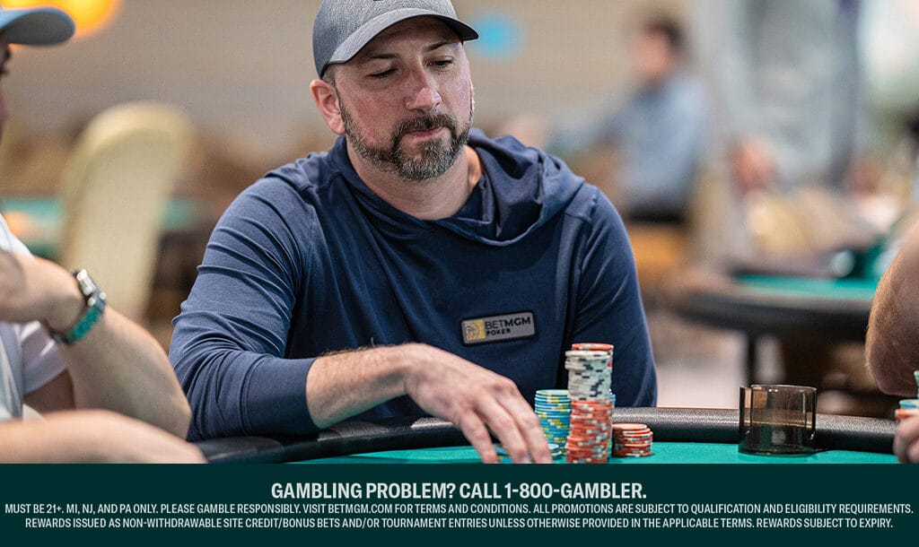 A man wearing a gray cap and blue hoodie with a BetMGM Poker logo sits at a poker table, stacking colorful chips. Other players and tables are blurred in the background. A responsible gambling message is displayed at the bottom.