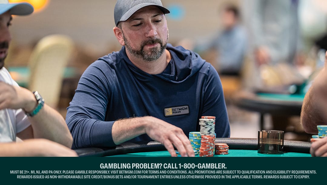 A man wearing a gray cap and blue hoodie with a BetMGM Poker logo sits at a poker table, stacking colorful chips. Other players and tables are blurred in the background. A responsible gambling message is displayed at the bottom.