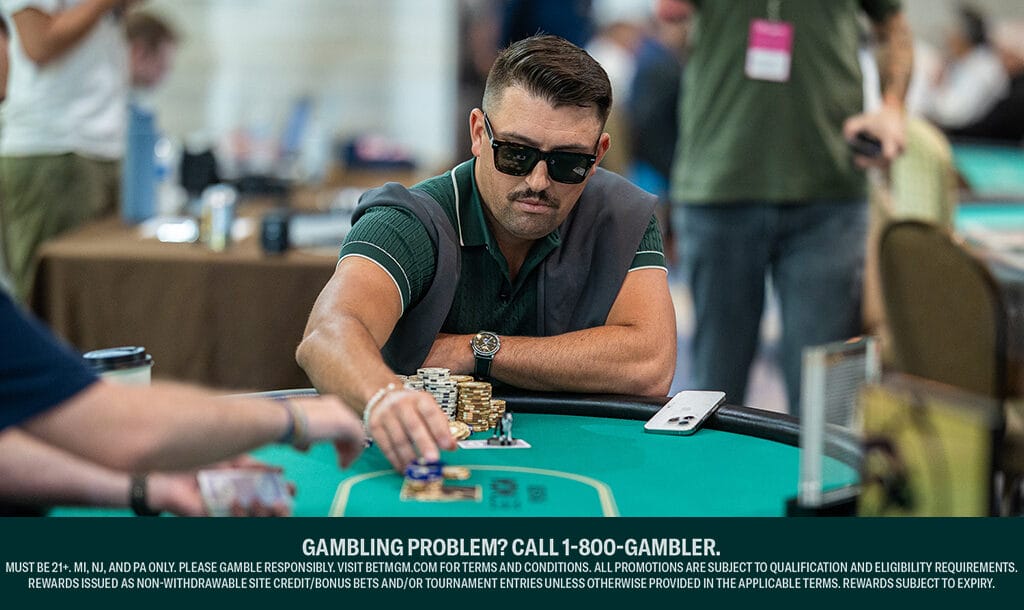 A man wearing sunglasses and a green shirt with a sweater draped over his shoulders plays poker at a casino table, reaching forward to move chips. Several stacks of chips and playing cards are on the table, and other players are partially visible in the background. A “GAMBLING PROBLEM? CALL 1-800-GAMBLER” disclaimer is displayed at the bottom.