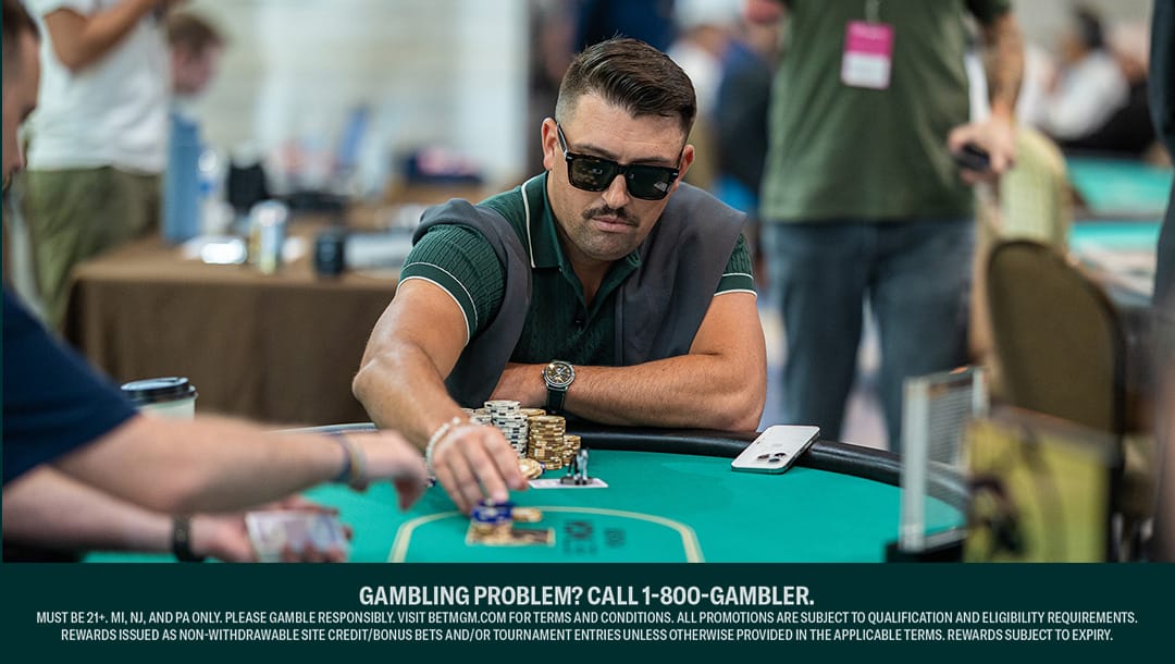 A man wearing sunglasses and a green shirt with a sweater draped over his shoulders plays poker at a casino table, reaching forward to move chips. Several stacks of chips and playing cards are on the table, and other players are partially visible in the background. A “GAMBLING PROBLEM? CALL 1-800-GAMBLER” disclaimer is displayed at the bottom.