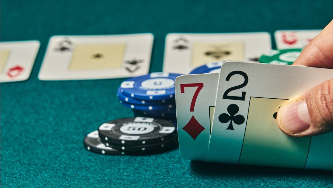 A poker player checks their hand. They see a 7 of diamonds and a 2 of clubs. There are poker chips and community poker cards in the background.