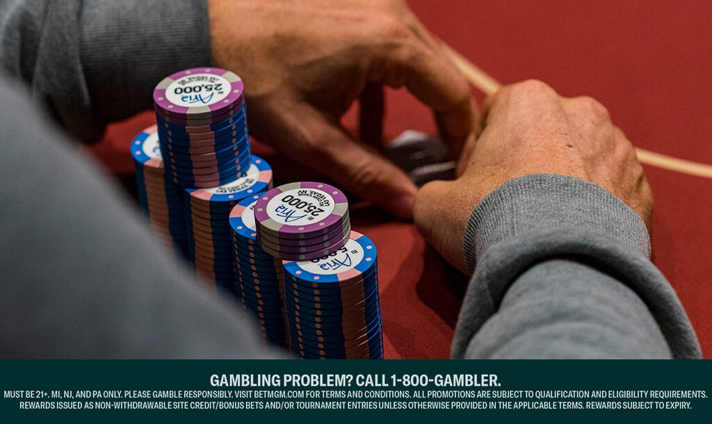 An over-the-shoulder view of a player at a poker table. There are multiple stacks of different colored poker chips from the Aria in front of them. Their hands are covering their cards.