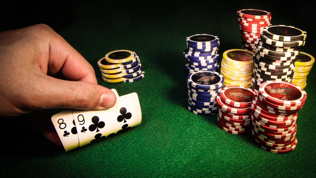 A player peeks at their hole cards and sees an eight and a nine of clubs. These cards are on a green poker table next to stacks of poker chips.